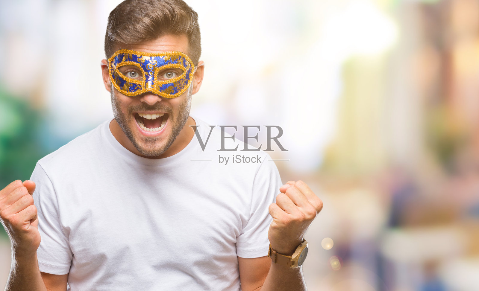 年轻英俊的男子戴着狂欢节面具，在孤立的背景下，举起双臂，睁开眼睛庆祝成功的惊喜和惊奇。赢家的概念。照片摄影图片