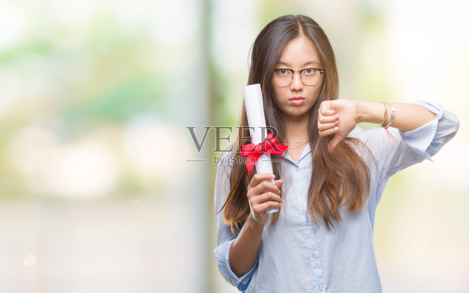 年轻的亚洲女性持有学位，在孤立的背景下，愤怒的脸，消极的迹象显示不喜欢拇指，拒绝的概念照片摄影图片