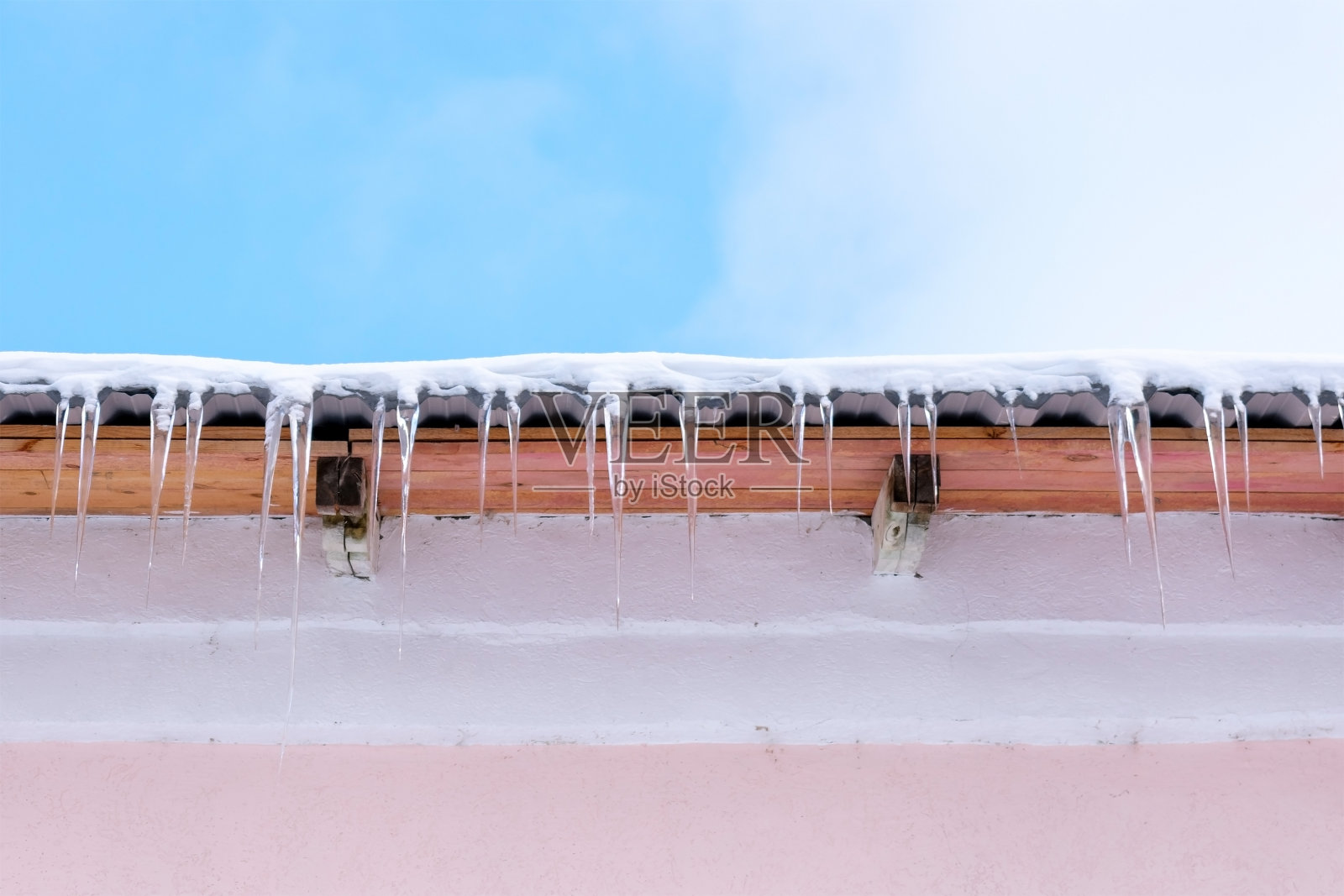 挂在粉红色建筑物屋顶上的冰柱。温暖晴朗的天气。照片摄影图片