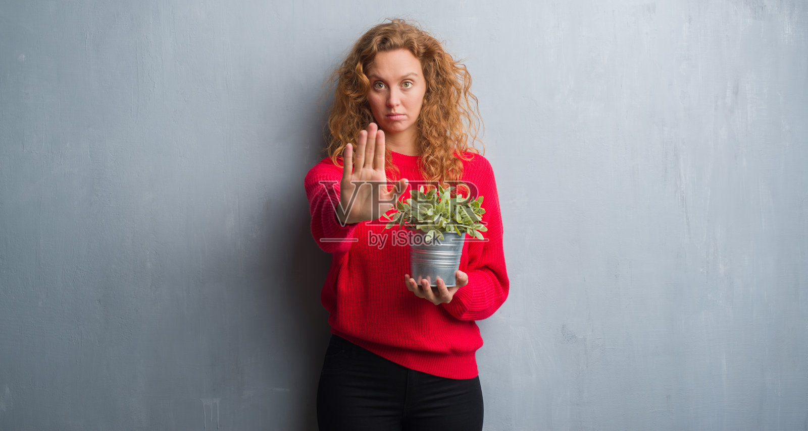 年轻的红头发的女人在灰色的垃圾墙拿着花盆与开放的手做停止标志与严肃和自信的表情，防御手势照片摄影图片