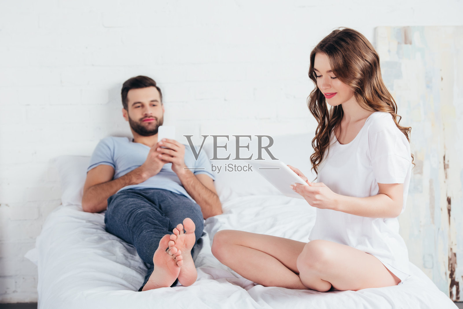 女人在用平板电脑，而男人躺在床上玩智能手机照片摄影图片