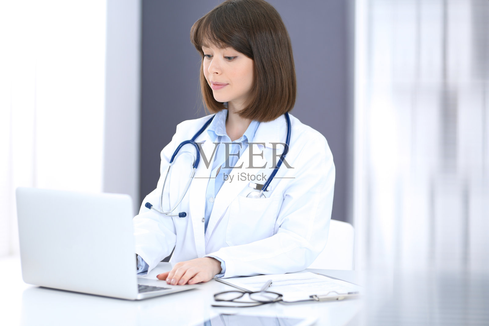 医生在笔记本电脑上打字，而坐在白色的桌子在医院办公室。医生在工作中。医疗保健理念照片摄影图片