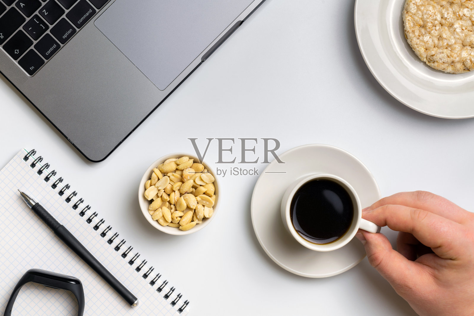 在工作休息时间吃健康的零食。运动员一边吃着花生，一边喝着咖啡，旁边放着笔记本电脑，健身追踪器和笔记本电脑。白色有组织的桌子。照片摄影图片