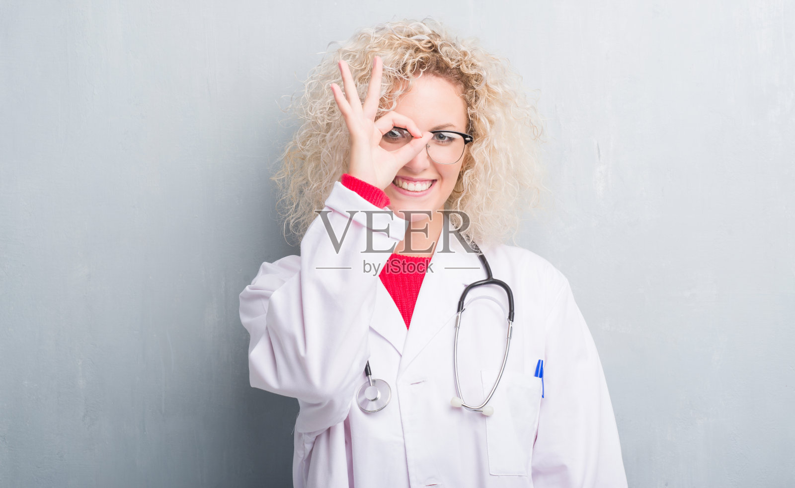 年轻的金发医生女人在垃圾灰墙做手势与手微笑，眼睛透过手指与快乐的脸。照片摄影图片