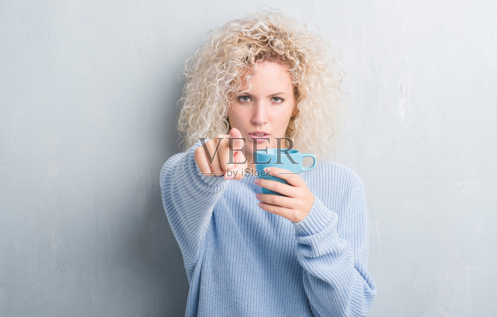 年轻的金发女人越过肮脏的灰色墙壁，喝着一杯咖啡，手指指向镜头和你，手势，正面自信的手势照片摄影图片