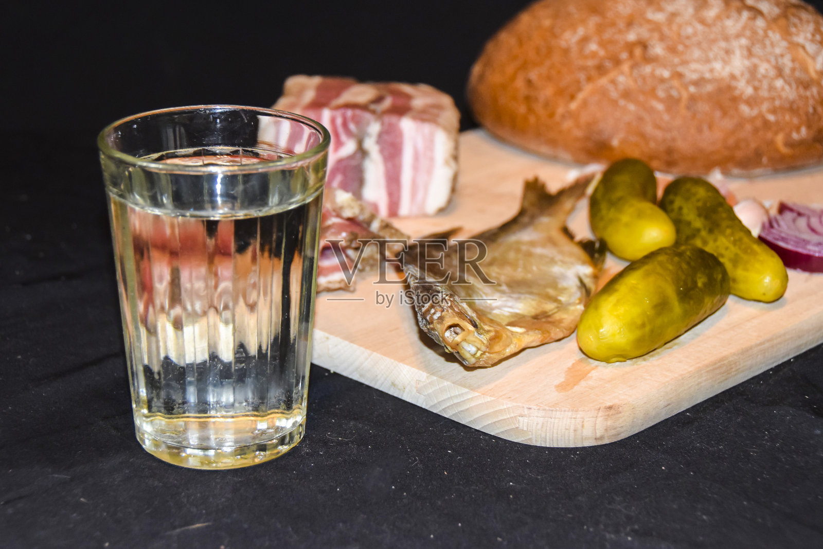 一杯俄罗斯伏特加和开胃菜沙罗鲷鱼、腌黄瓜、洋葱、大蒜面包放在一块木板上，背景是黑色照片摄影图片
