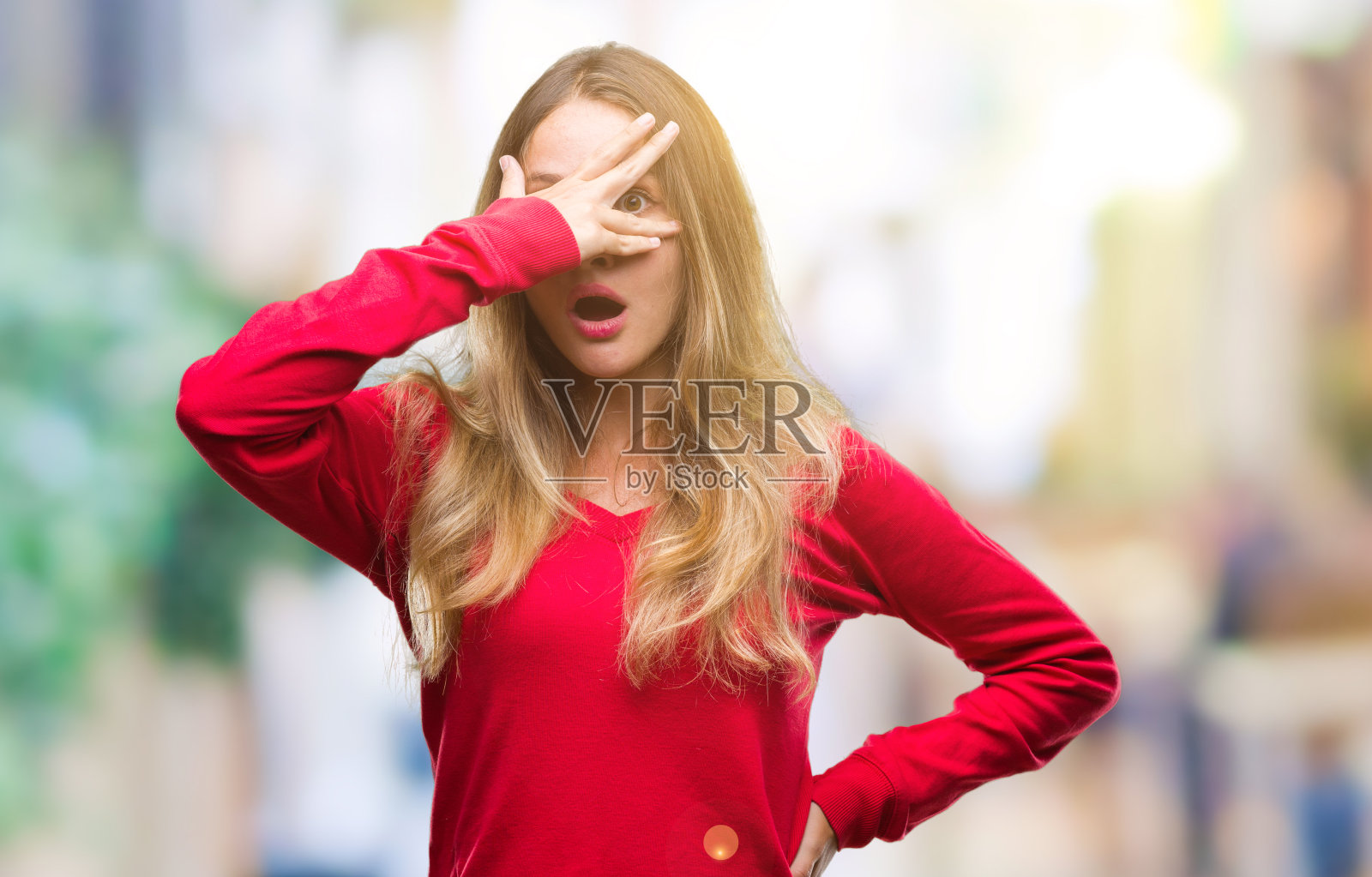 年轻美丽的金发女人穿着红色毛衣在孤立的背景中窥视在震惊覆盖的脸和眼睛用手，透过手指看着尴尬的表情。照片摄影图片