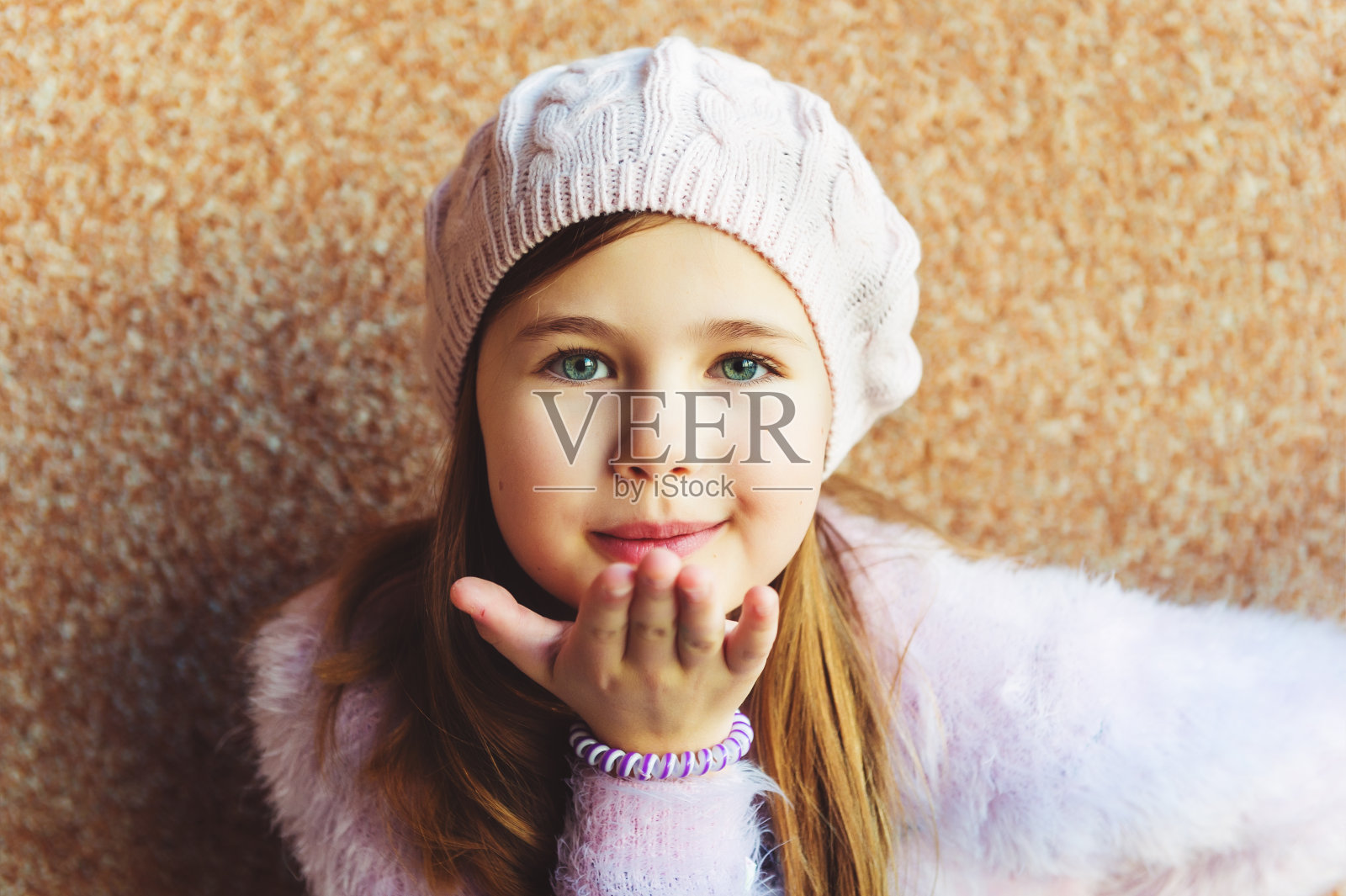 近距离的肖像可爱的9岁小女孩与蓝色的眼睛，戴粉红色的帽子，站在棕色的背景。一个孩子用她的手送一个吻照片摄影图片