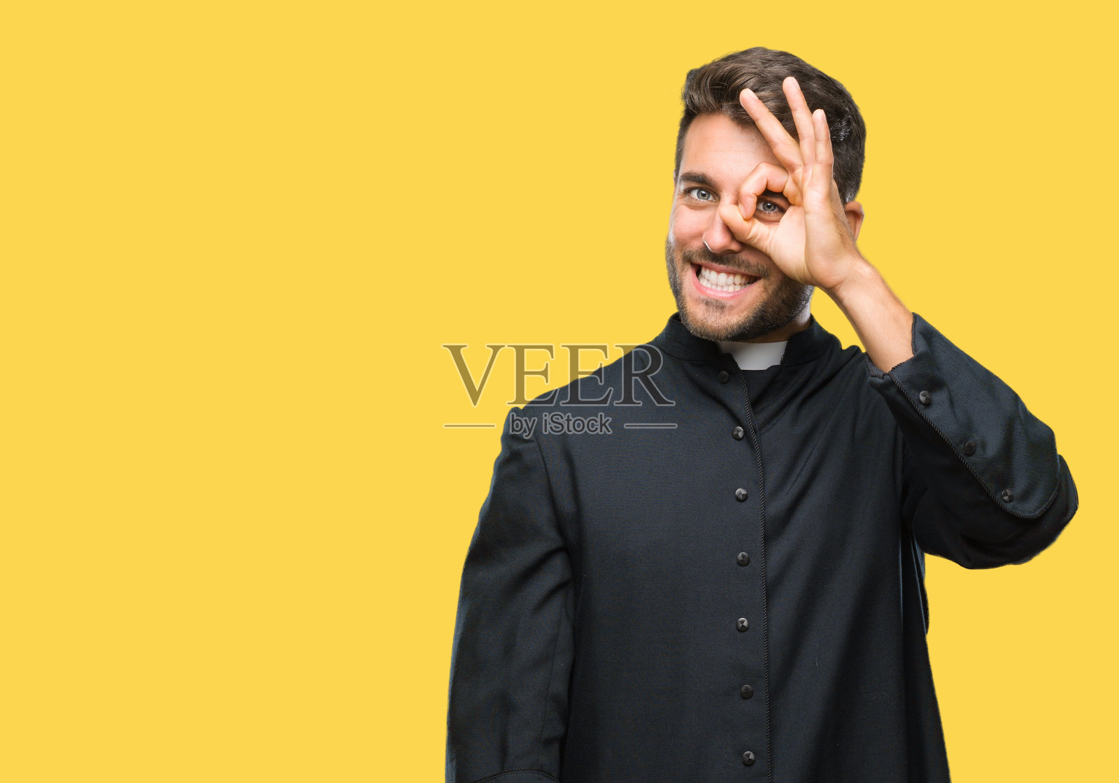 年轻的天主教牧师男人在孤立的背景做ok手势与手微笑，眼睛透过手指与幸福的脸。照片摄影图片