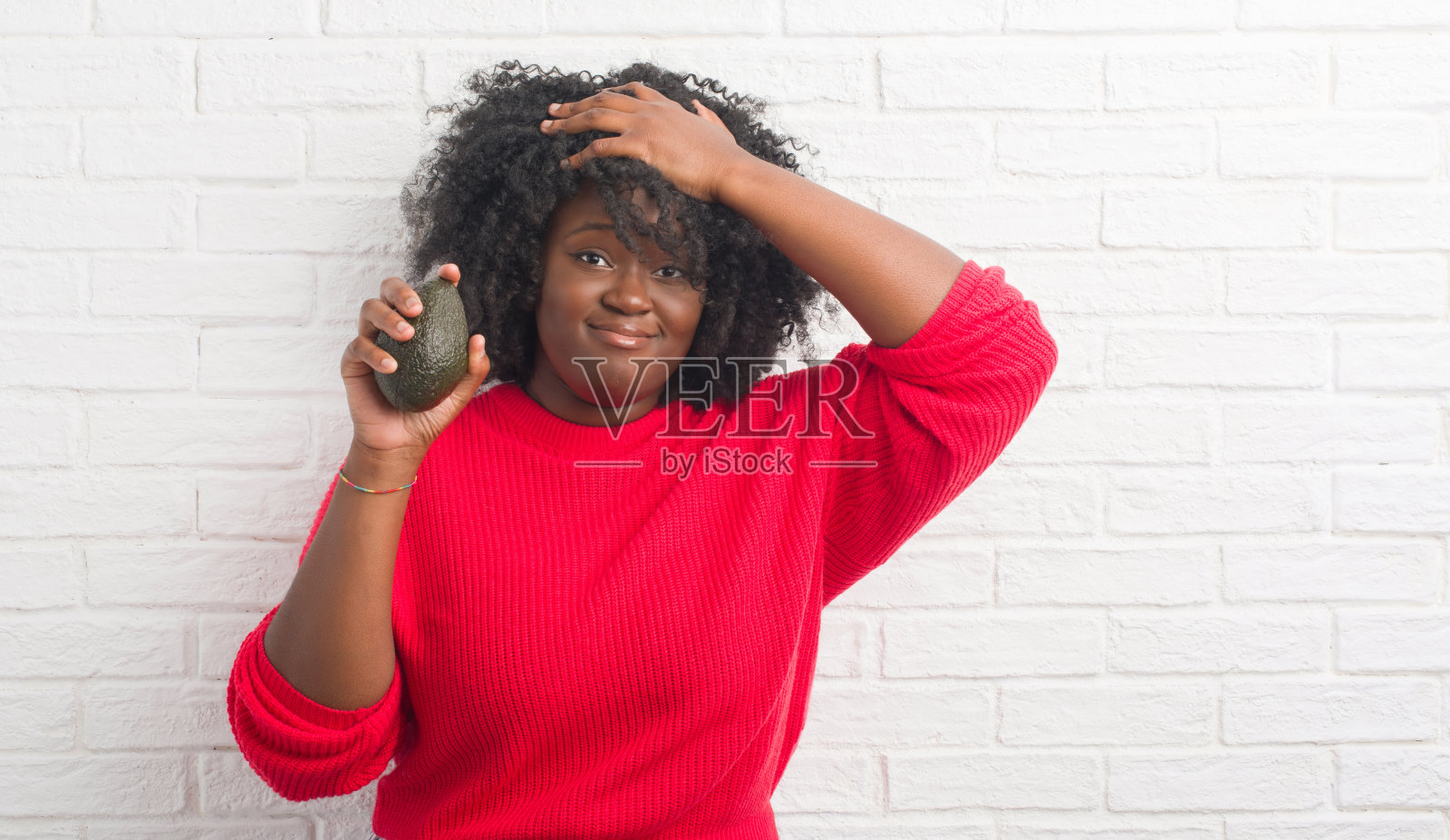 年轻的非洲裔美国妇女越过白砖墙吃牛油果强调用手顶着头，震惊的羞愧和惊讶的脸，愤怒和沮丧。为错误而恐惧和不安。照片摄影图片