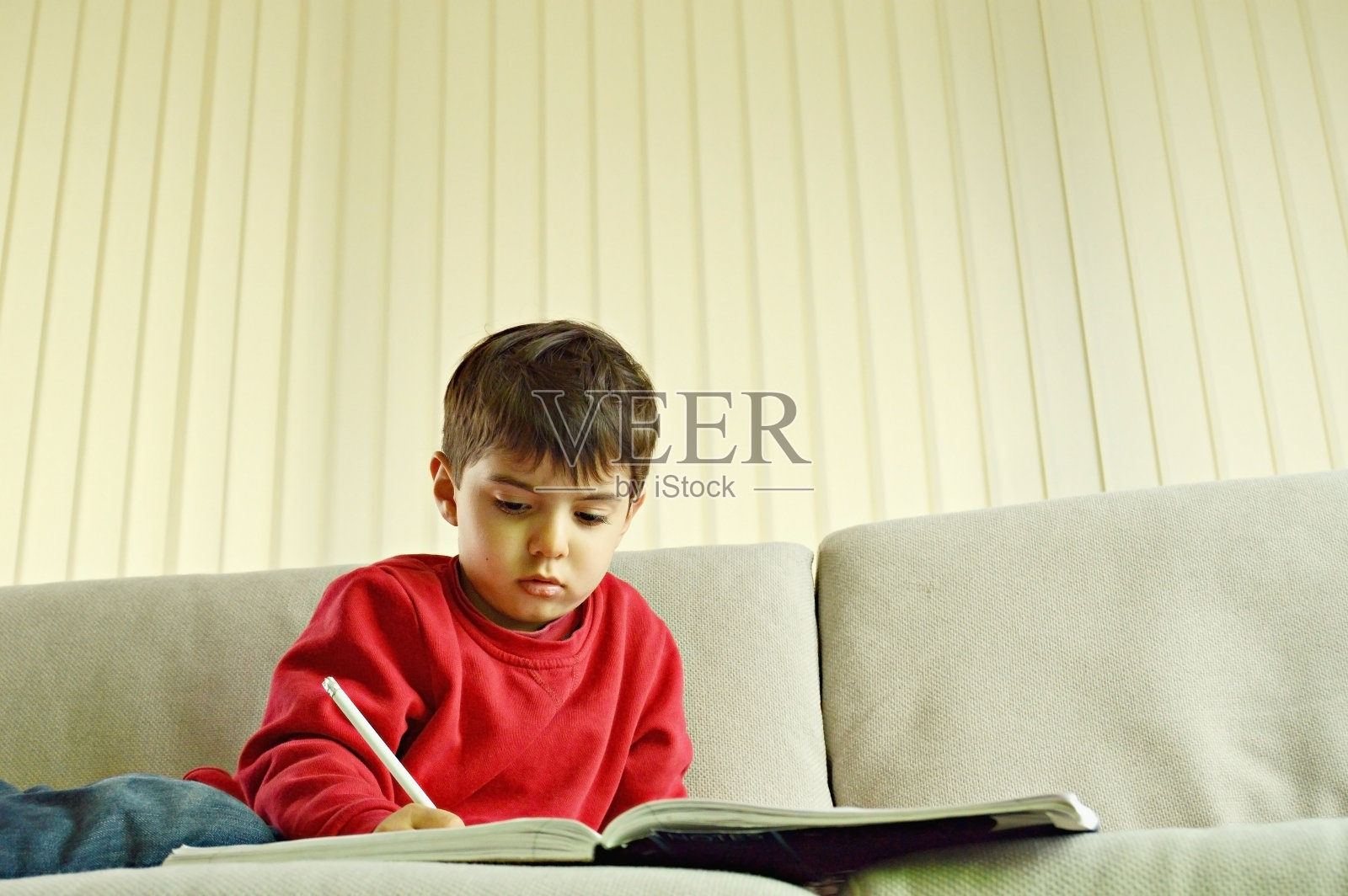 一个可爱的学前儿童穿着红色的套头衫和蓝色的牛仔裤，坐在一个现代客厅的白色沙发上用白色的铅笔在笔记本上写东西照片摄影图片