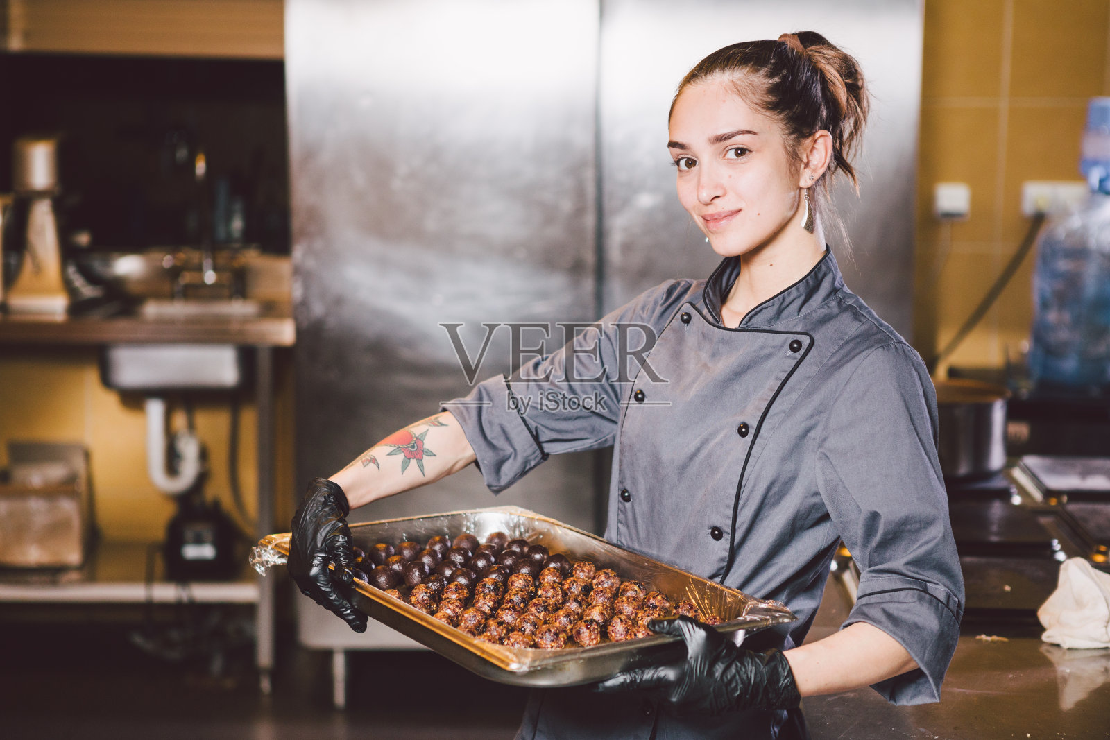 主修专业及烘焙糕点。一名纹身是糕点师的年轻白种女子，穿着黑手套和制服，在餐厅厨房准备圆形巧克力糖果、手工松露照片摄影图片