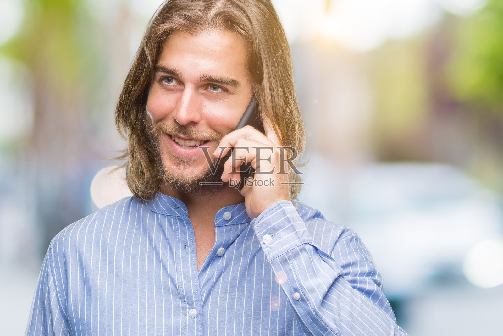 年轻英俊的男人长头发在孤立的背景下讲电话与一个快乐的脸站着，微笑着自信的微笑显示牙齿照片摄影图片