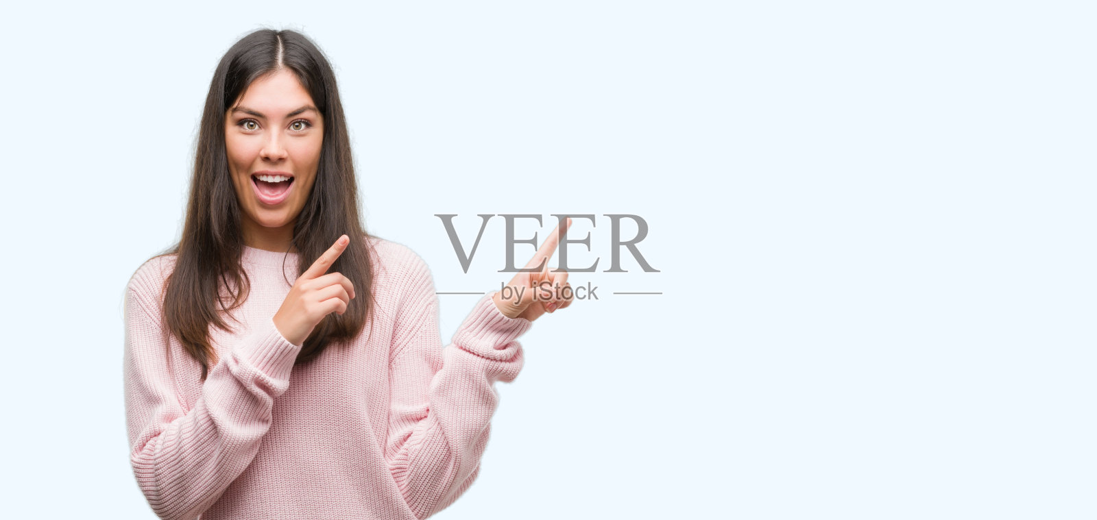 年轻美丽的西班牙妇女穿着一件毛衣，微笑着看着镜头，两只手和手指指向一边。照片摄影图片