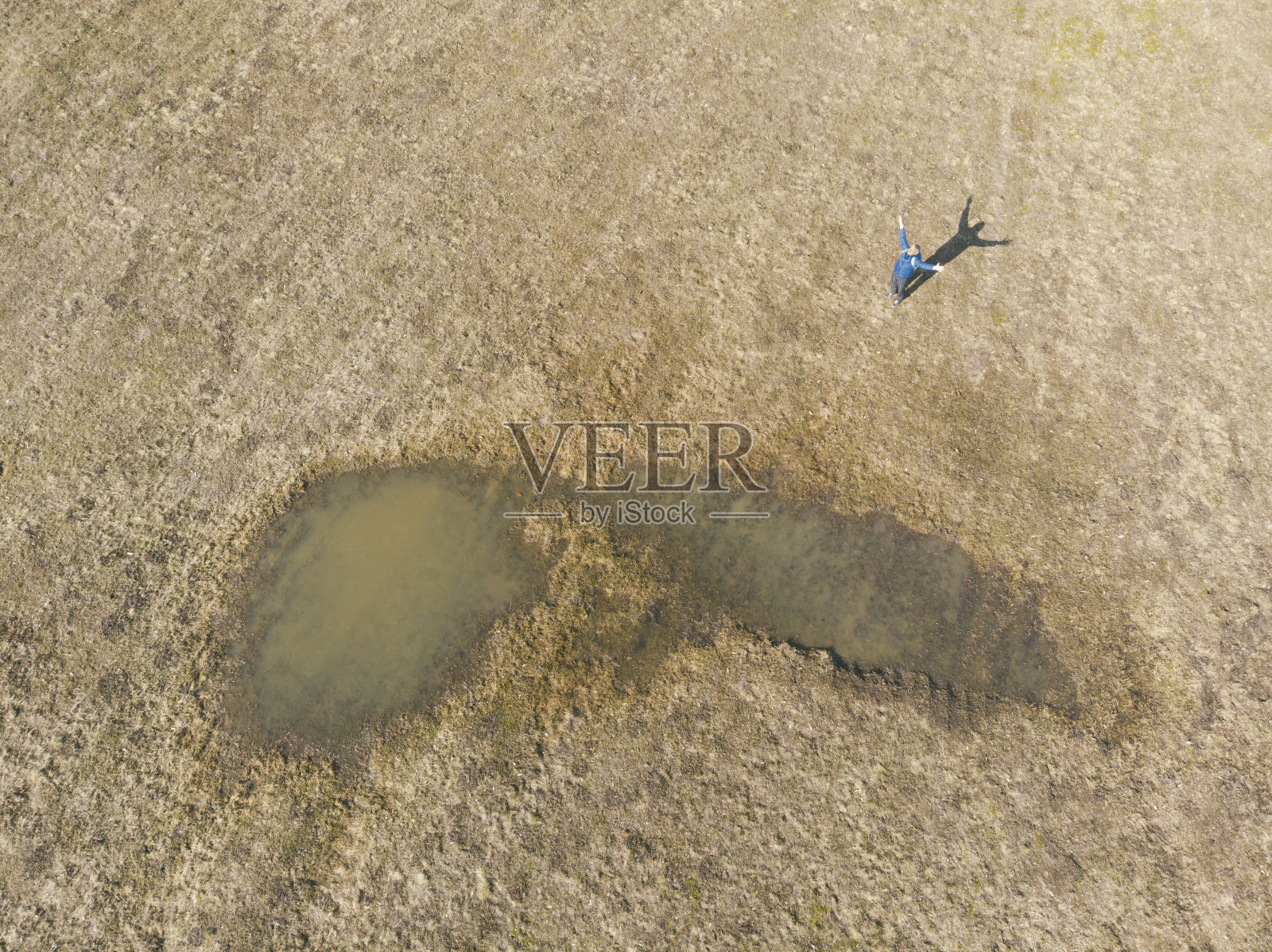 一名成年男子伸开双臂，在冬末阳光明媚的日子里，在小水塘旁边的干草地上徒步旅行。鸟瞰图。照片摄影图片