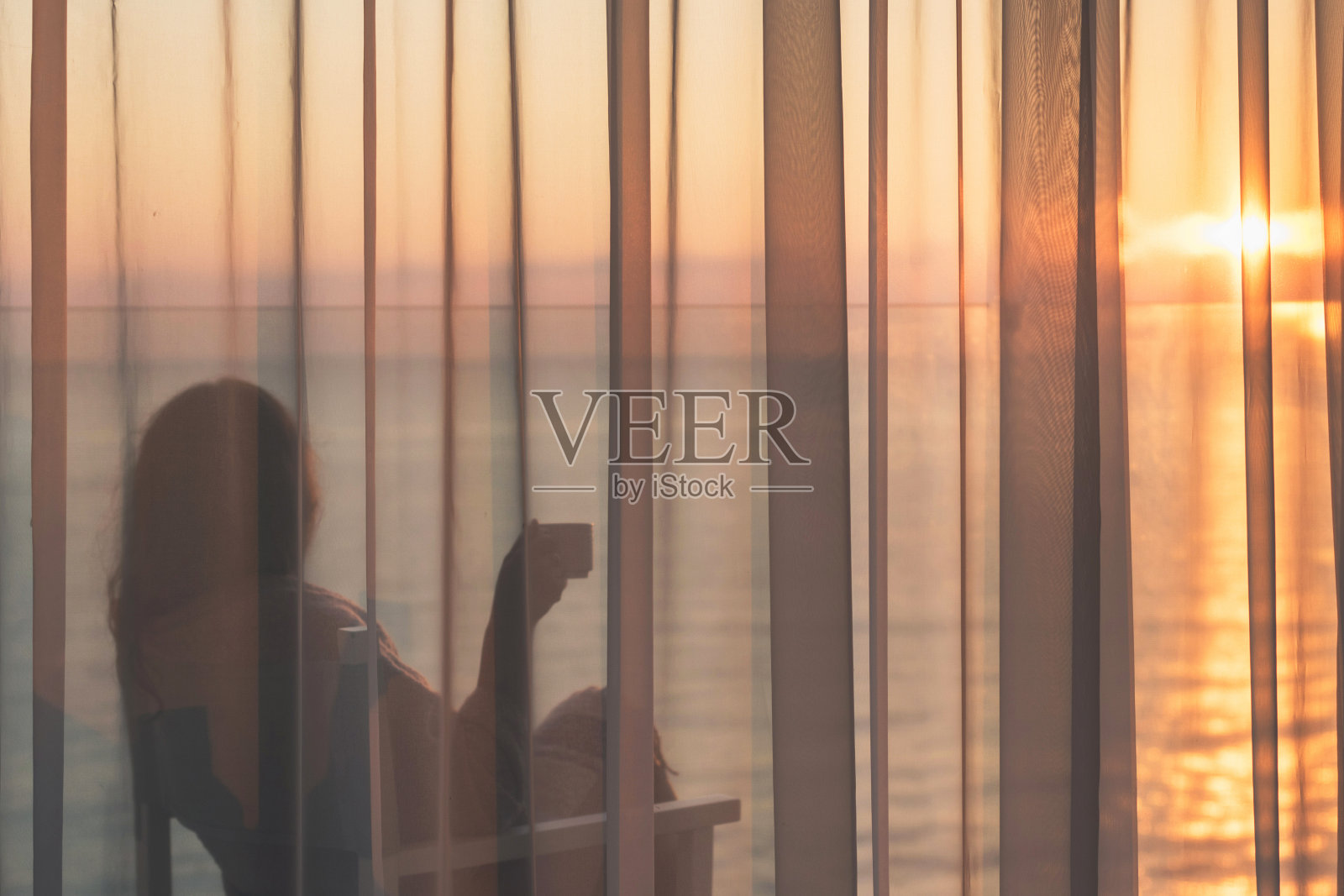 清晨日出时分，美女裹着毯子坐在玻璃阳台上的透明窗帘后，端着一杯咖啡坐在椅子上。海景照片摄影图片