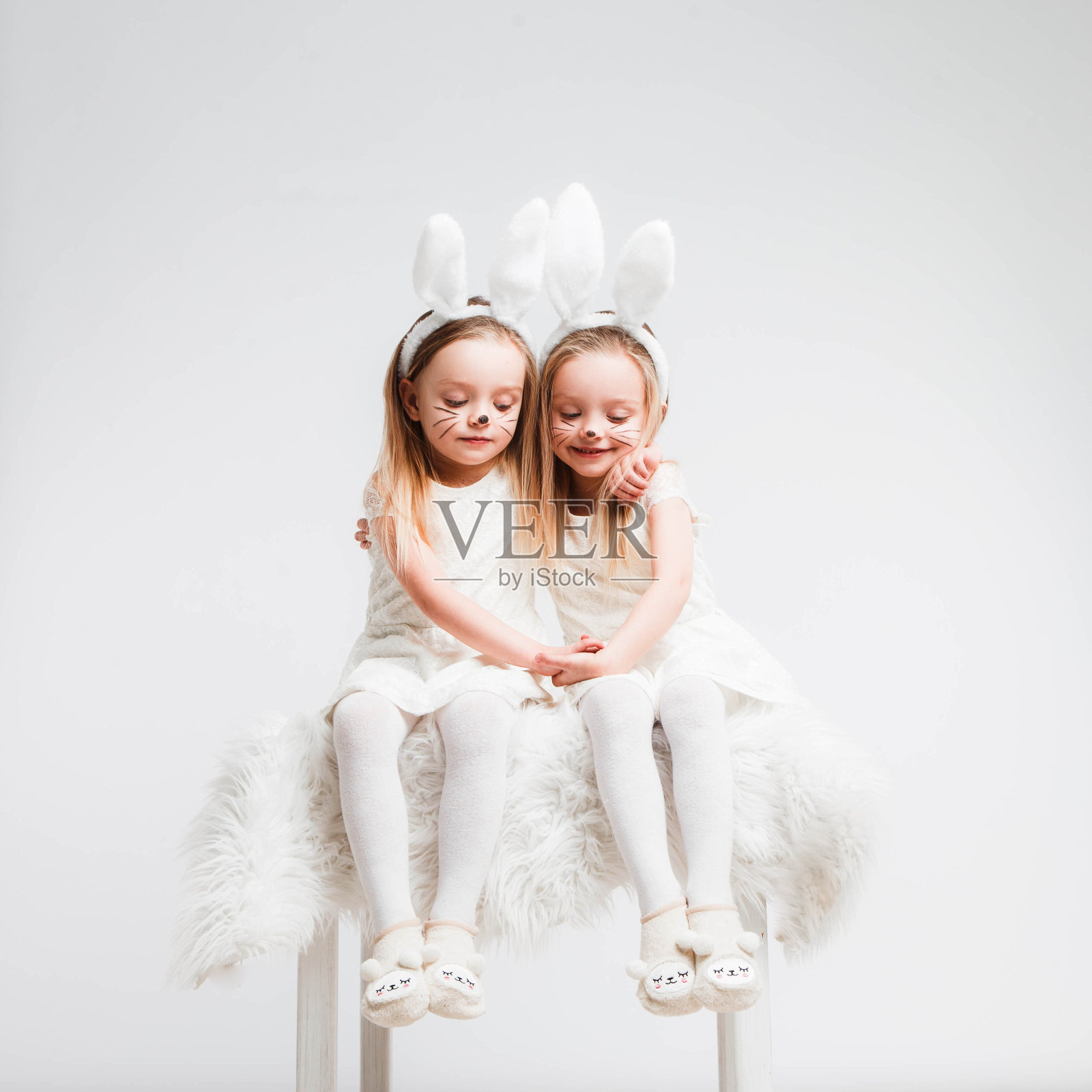 一对金发的双胞胎，穿着白裙子，长着兔耳朵。灰色背景上的工作室照片。孩子们庆祝复活节。照片摄影图片