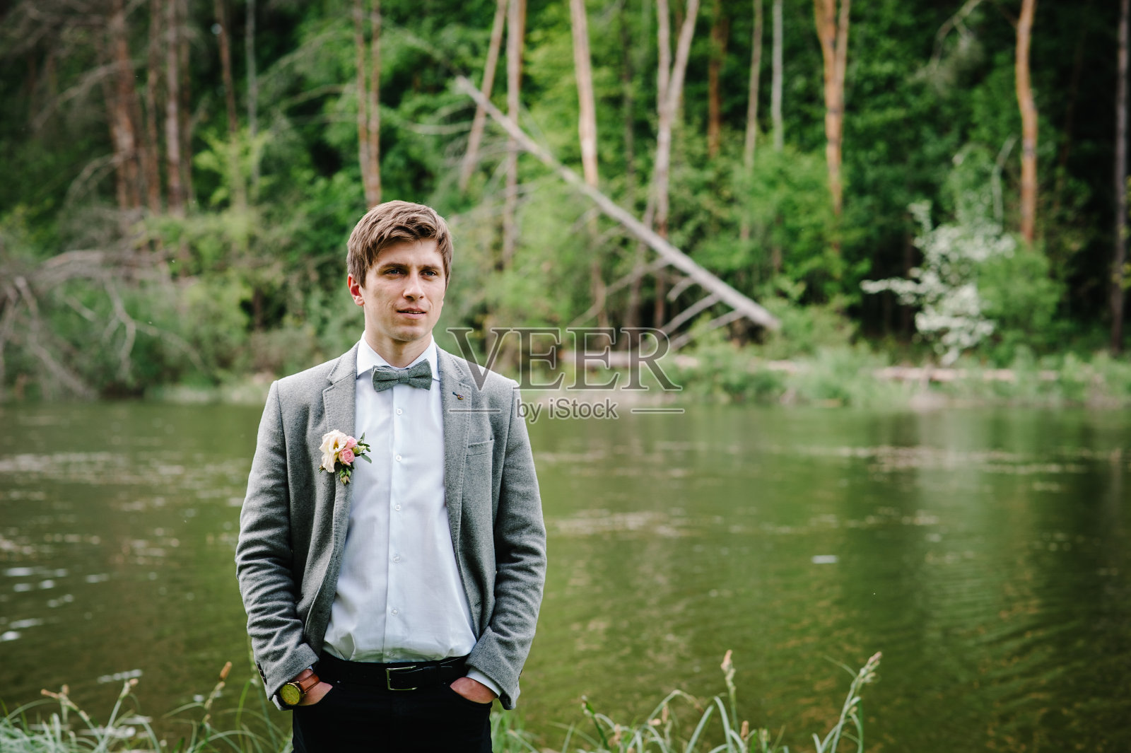 照片中的新郎身穿西装，系着领结，夹克上有纽扣或纽扣孔，背景是河边或湖边的自然森林。照片摄影图片