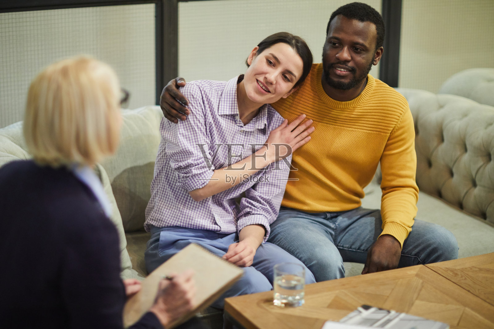 积极年轻的跨种族夫妇在休闲服装坐在沙发上拥抱，而谈话的房地产经纪人和分享他的要求照片摄影图片