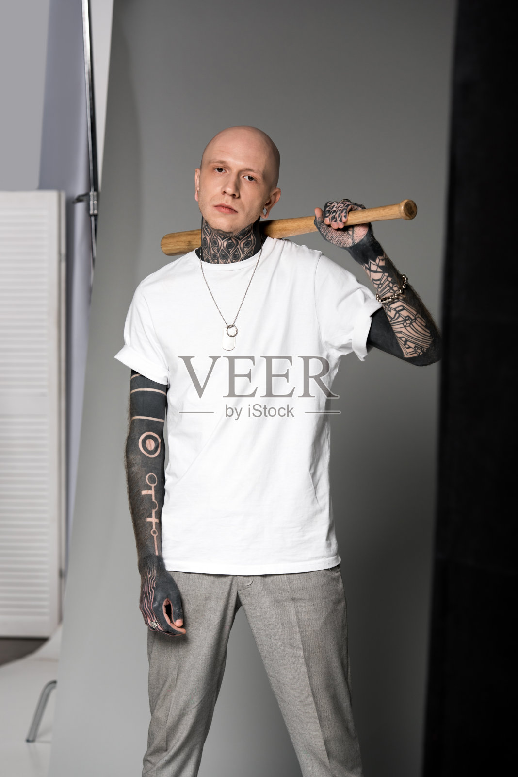 一名身穿白色t恤的光头纹身男子，肩上扛着棒球棒，站在摄影棚里看着摄像机照片摄影图片