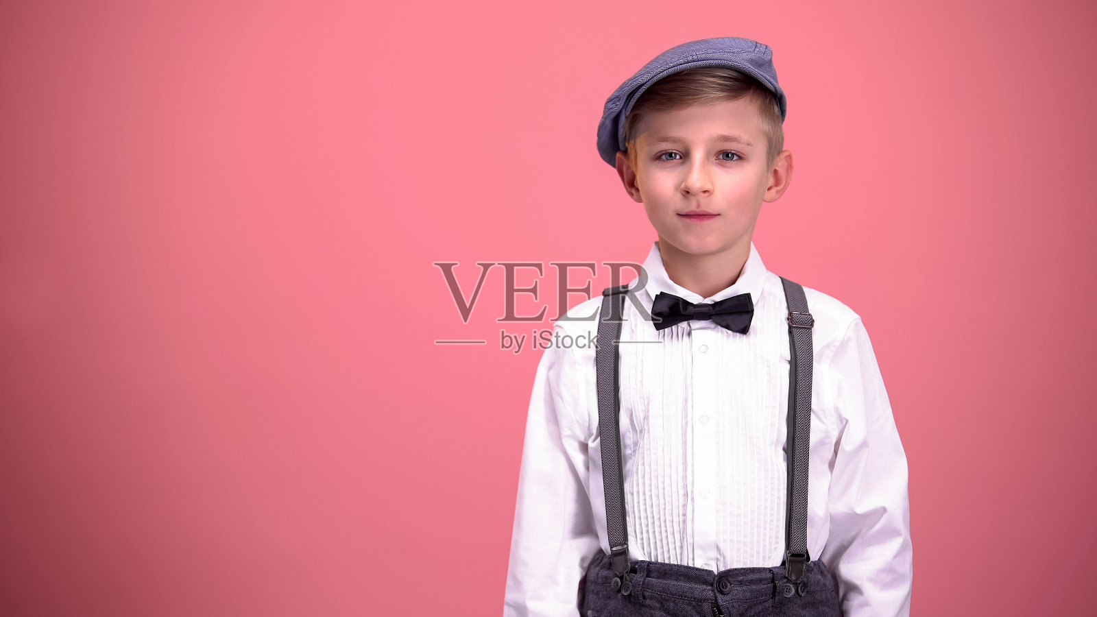 穿着复古衣服的小男孩看着镜头，在粉红色的背景上孤立无援照片摄影图片
