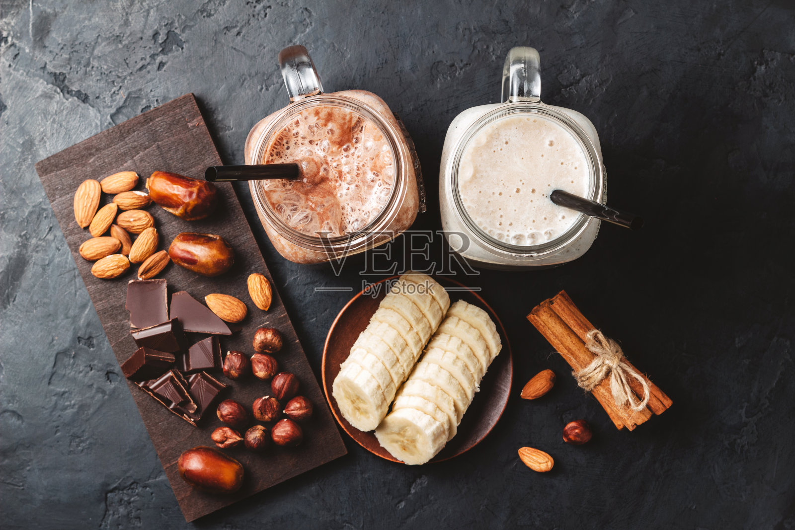 香蕉和巧克力冰沙在两个罐子，蛋白质奶昔，坚果，巧克力块，香蕉片，枣，肉桂棒，健康的饮食在黑暗的背景，俯视图照片摄影图片