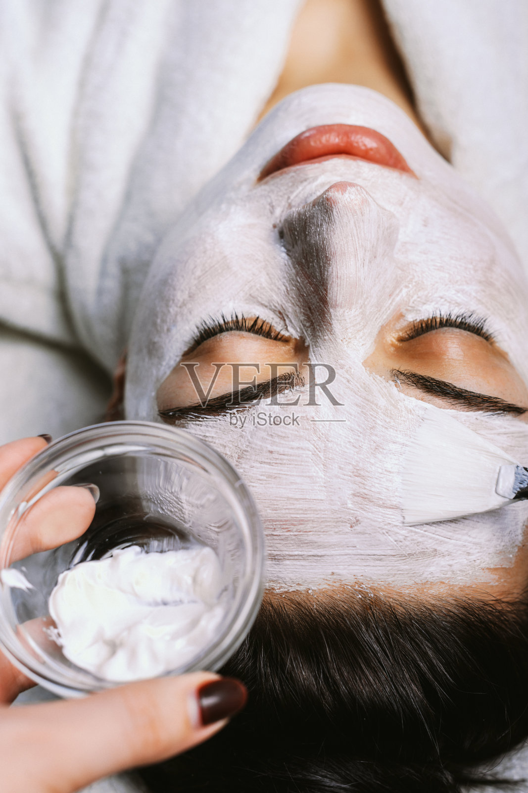 近距离观看一个女人的脸坐着闭上眼睛微笑，而她的脸上正在应用一个白色的面具在一个水疗中心。可爱的女性照顾她的皮肤在水疗沙龙。照片摄影图片