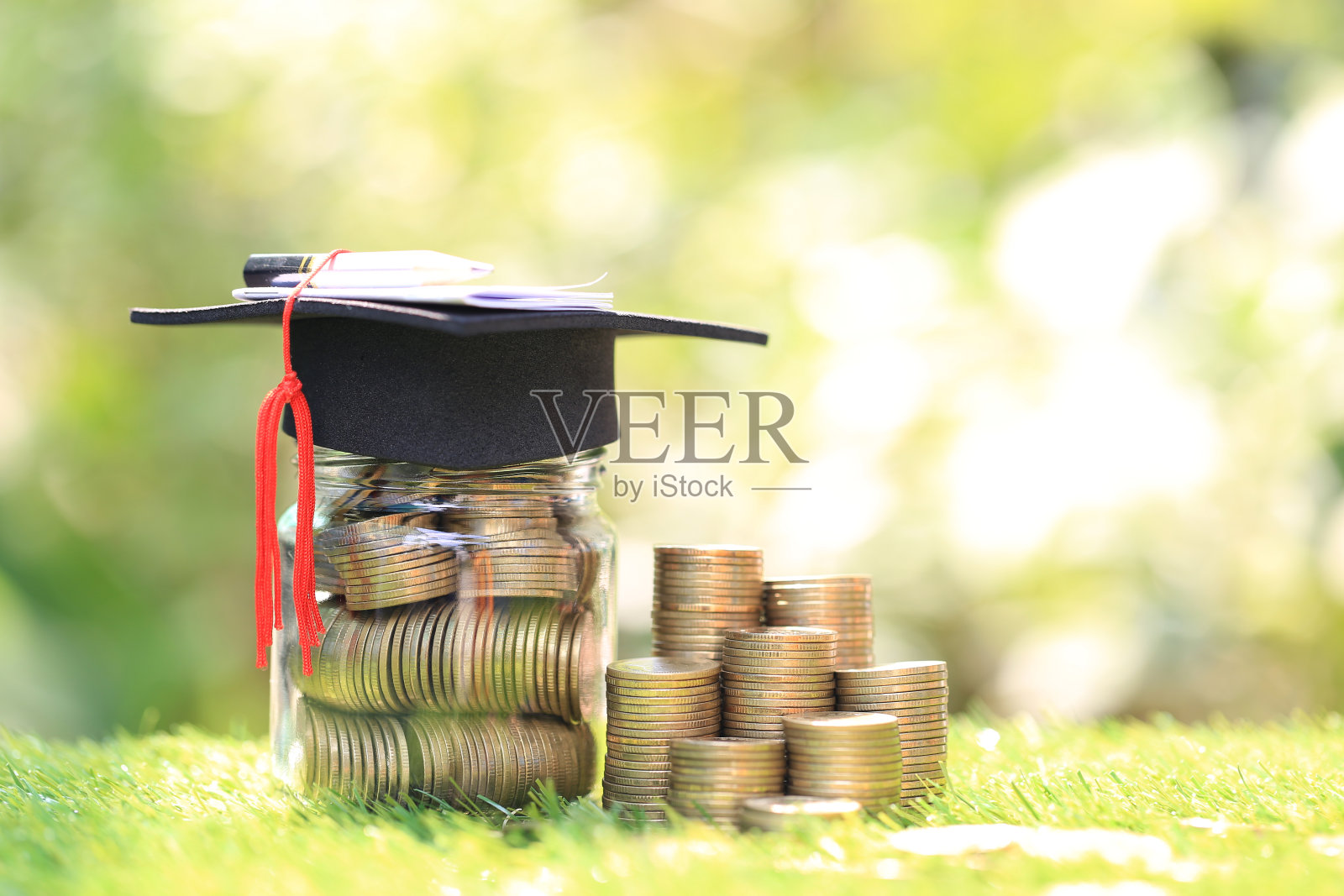 毕业帽上的玻璃瓶上有一叠硬币，钱以自然绿色为背景，省钱为教育理念照片摄影图片