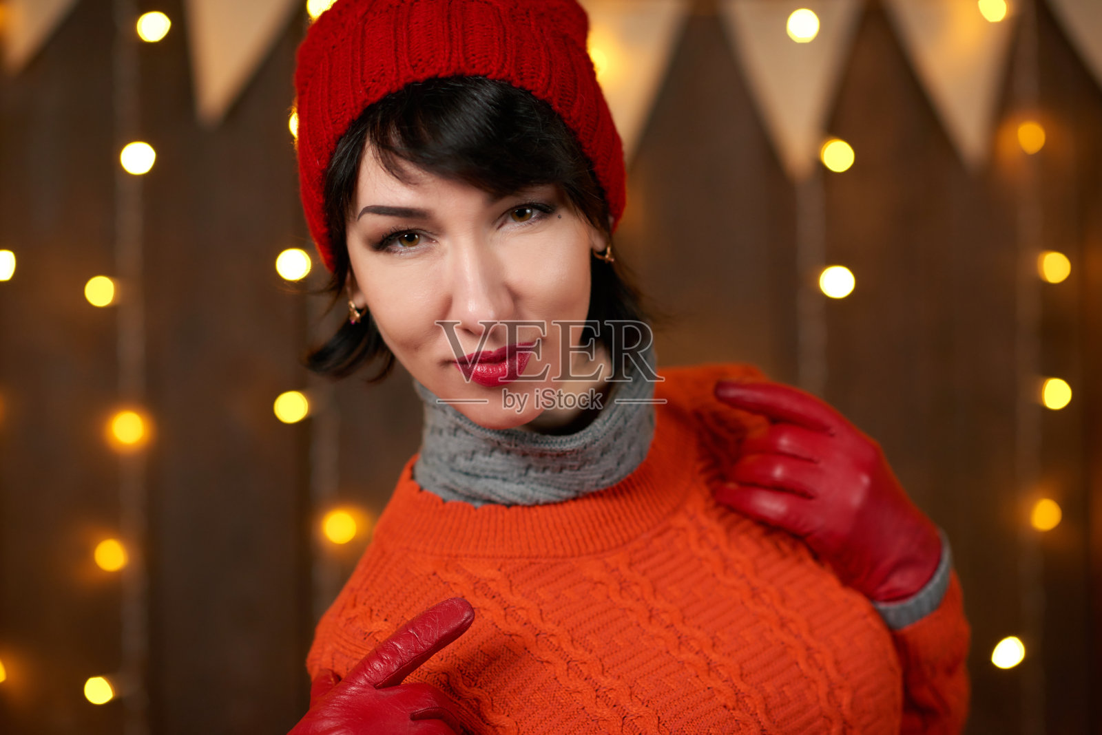 美丽的女人是摆着深色木制背景，圣诞灯和旗帜，节日的概念。穿着红色的针织帽和毛衣。照片摄影图片