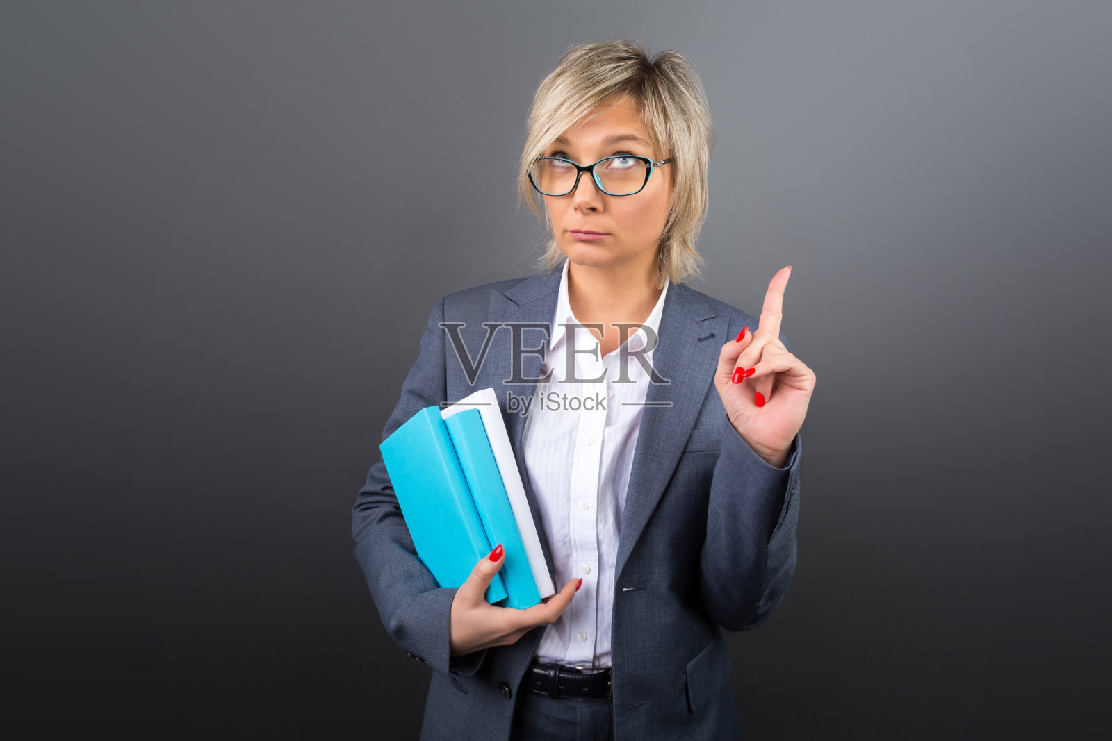 一个40多岁的金发女人的肖像，女商人，教师，教练身穿灰色西装，戴着绿松石眼镜，在灰色的背景下手里拿着书照片摄影图片