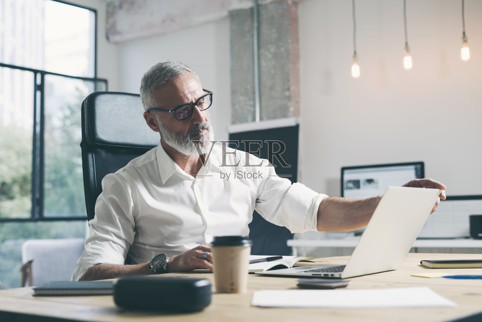 有魅力和自信的成年商人使用移动笔记本电脑，同时在现代共同工作办公室的木桌上工作。照片摄影图片