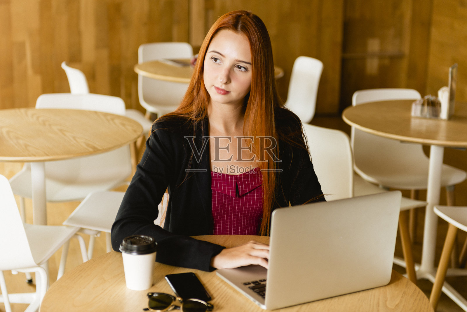 漂亮的红发模特在用电脑。桌子上放着手机太阳镜和咖啡杯，背景模糊照片摄影图片