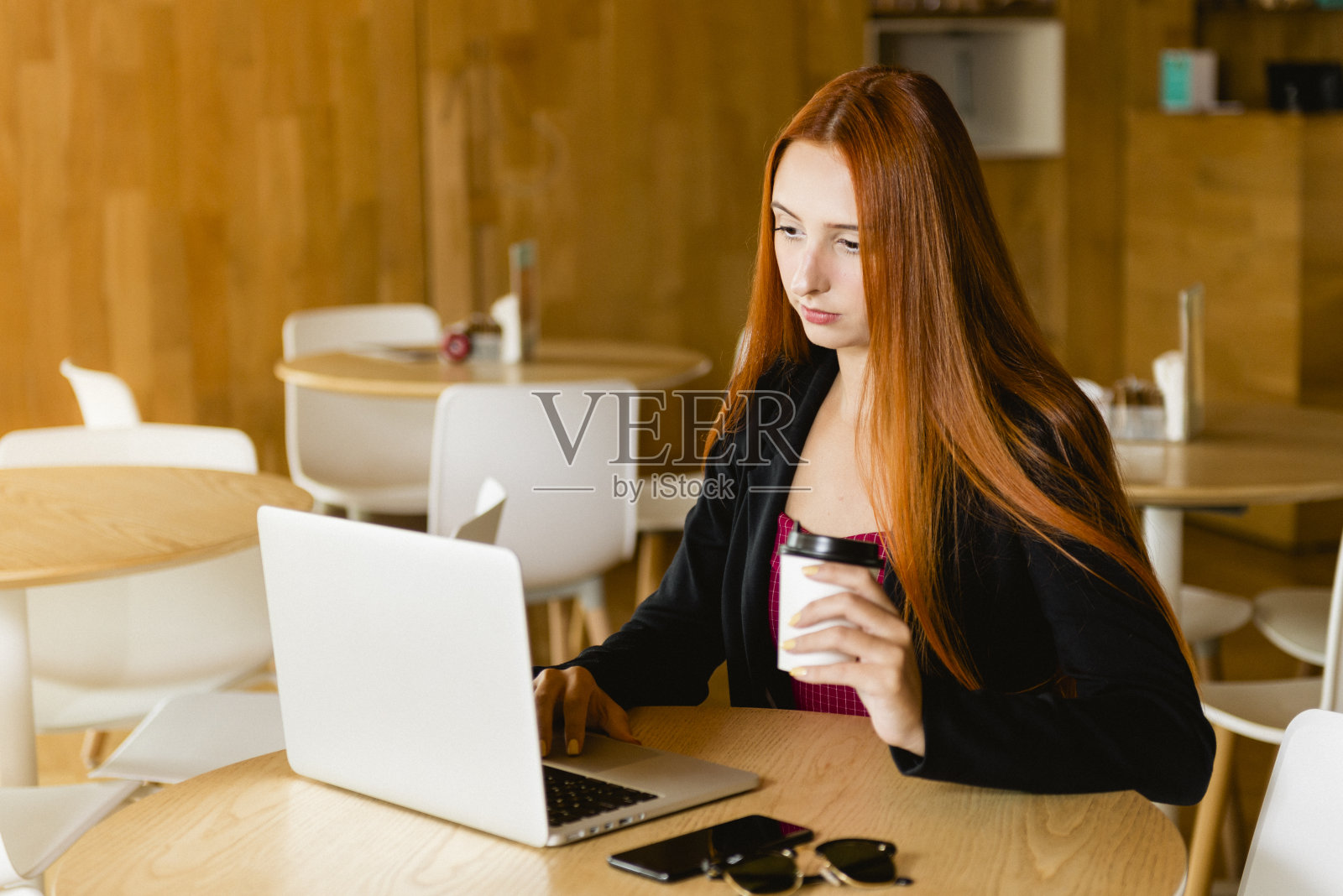 漂亮的红发模特拿着一杯咖啡用电脑。桌子上放着手机和墨镜，背景模糊照片摄影图片