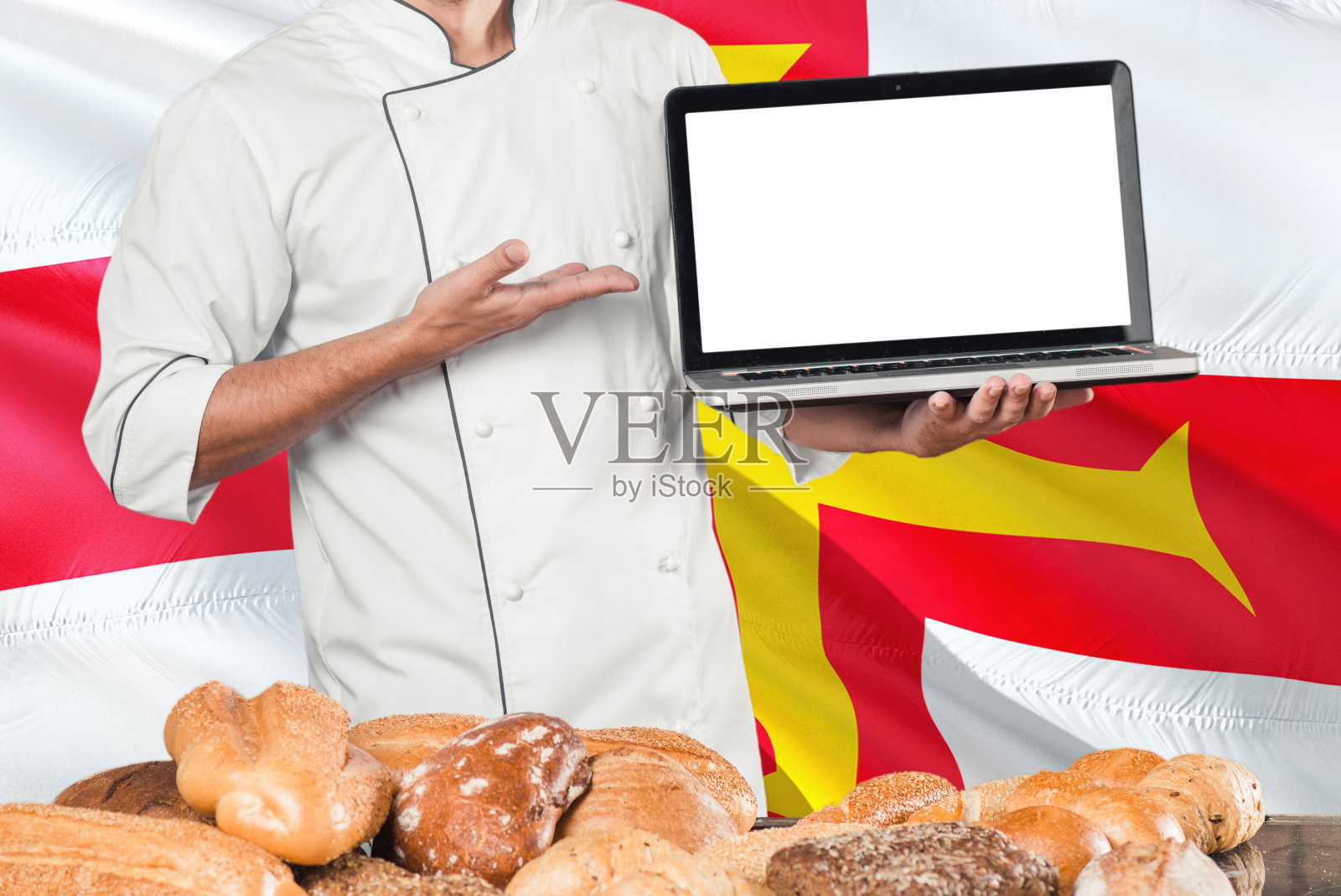 面包师拿着笔记本电脑在根西岛旗和面包的背景。厨师穿着制服指向空白屏幕为拷贝空间。照片摄影图片