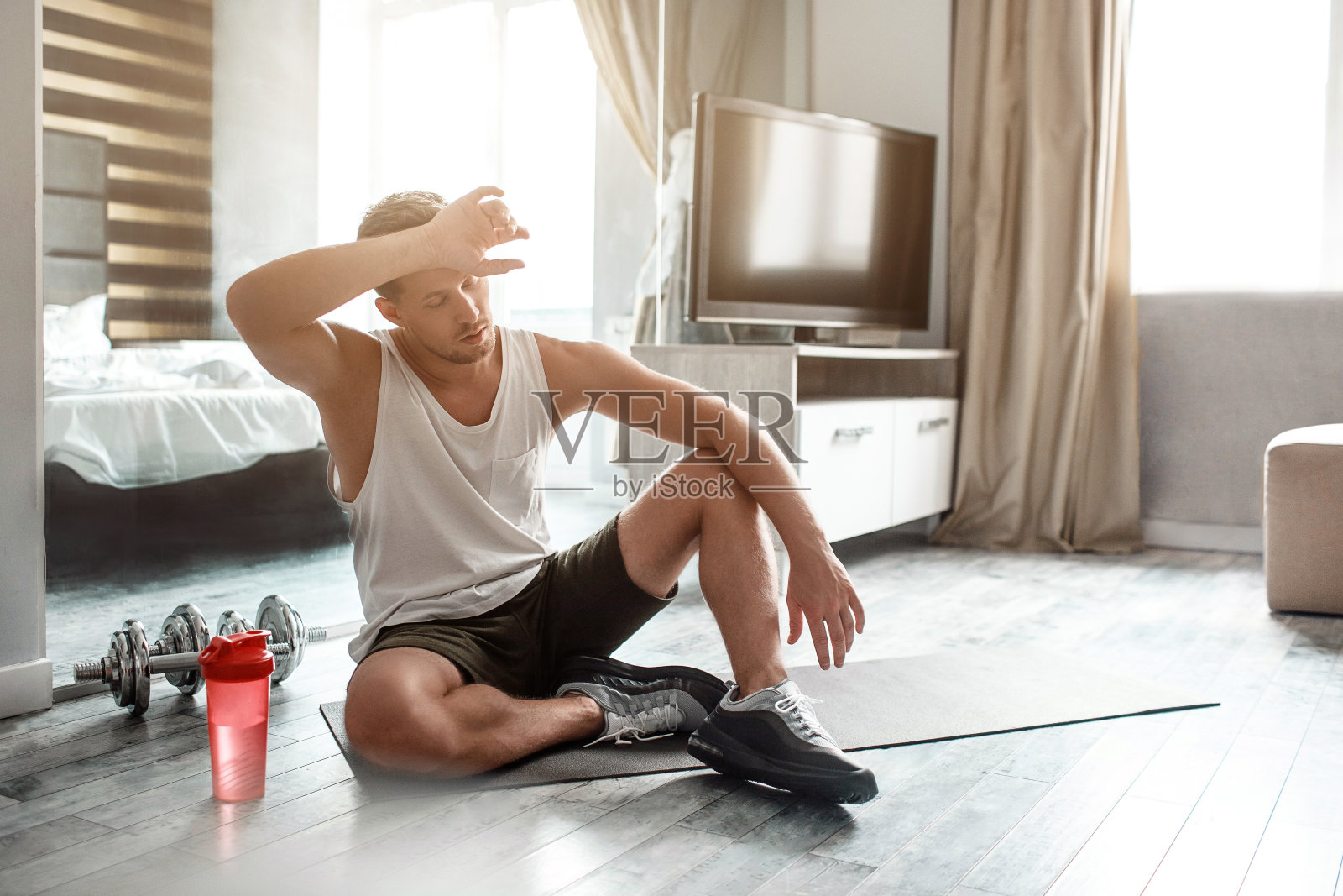 体格健壮的年轻男子在公寓里参加体育活动。精疲力竭的家伙在锻炼后。他坐在地板上，手放在额头上。照片摄影图片