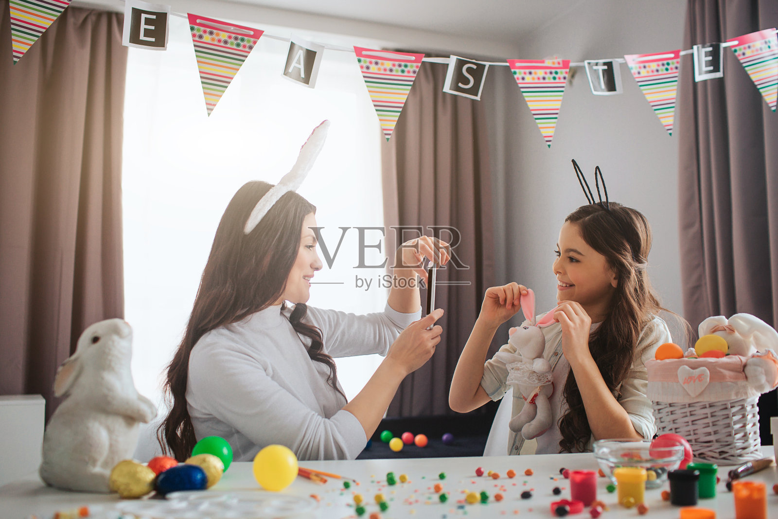 母亲和女儿准备复活节。年轻女子拍照的女孩与兔子玩具。女孩的姿势。他们戴着复活节兔子耳朵。人们坐在桌子旁的房间里。照片摄影图片
