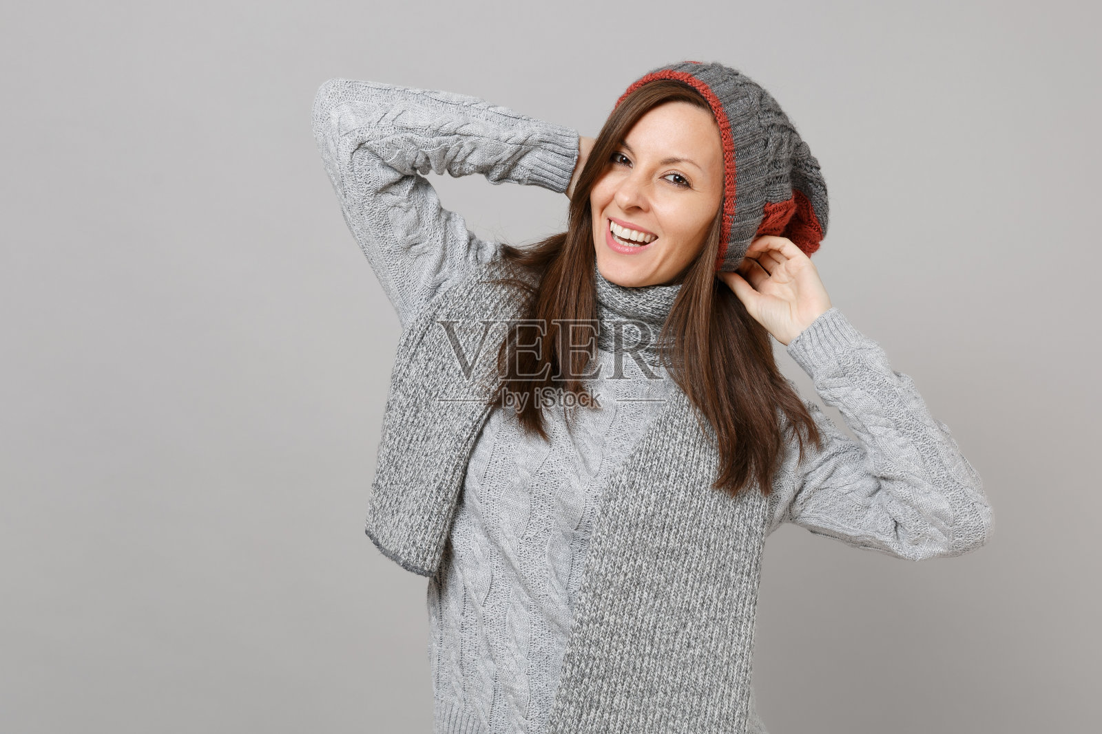 快乐的年轻女子在灰色毛衣，帽子和围巾把双手放在孤立在灰色背景的头在演播室。健康时尚的生活方式，真诚的情感，寒冷的季节理念。模拟拷贝空间。照片摄影图片