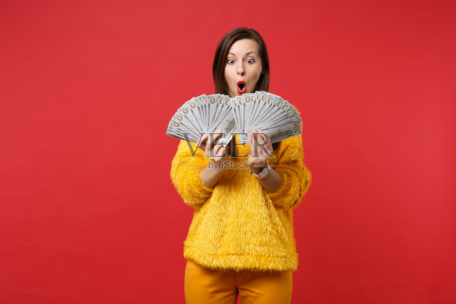 惊奇的年轻女子在皮草毛衣保持嘴大张着扇子的美元钞票现金钱孤立在红色的背景。人真诚情感生活理念。模拟拷贝空间。照片摄影图片