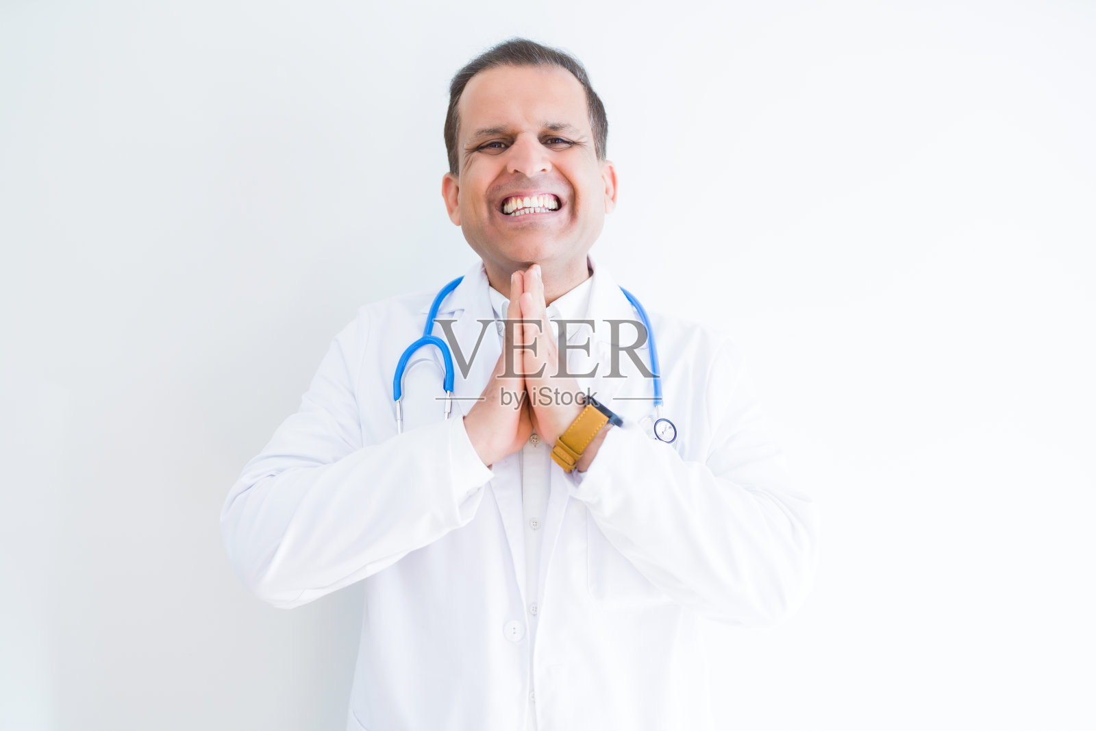 中年医生男子穿着听诊器和医疗外套在白色的背景祈祷与手一起请求原谅自信的微笑。照片摄影图片