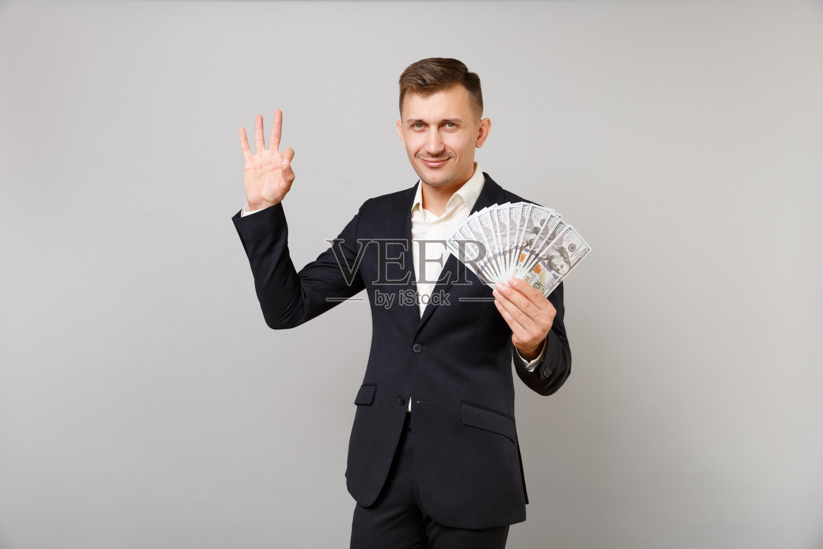 成功的年轻商人西装，显示OK手势，拿着一堆美元钞票现金孤立在灰色的背景。成就事业财富经营理念。模拟拷贝空间。照片摄影图片