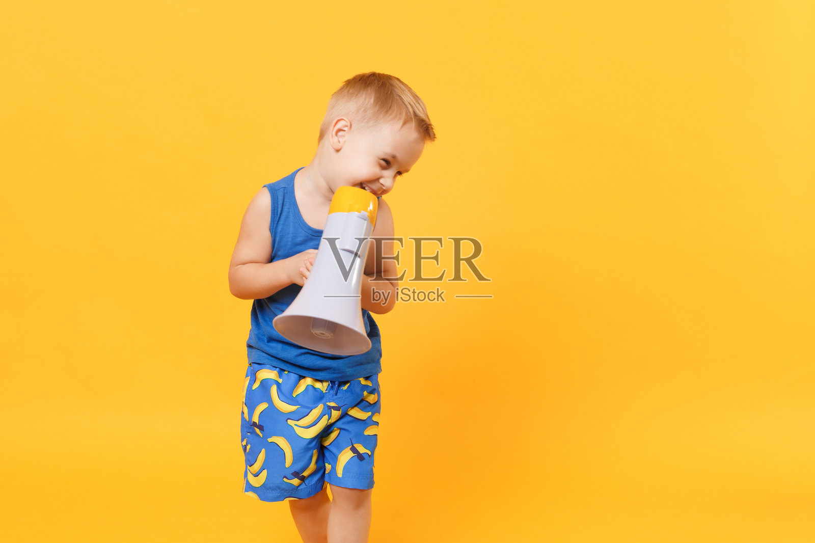 小男孩3-4岁，穿着蓝色的沙滩夏装，在扩音器里说话，孤立在明亮的橙黄色背景下的儿童摄影棚肖像。人们童年生活方式概念模拟复制空间。照片摄影图片
