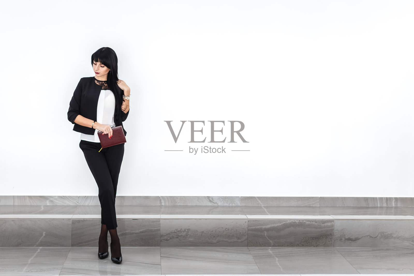 年轻、迷人、严肃的黑发女子，带着一本笔记本，穿着黑色的商务套装，靠墙站在办公室里。副本的空间。照片摄影图片