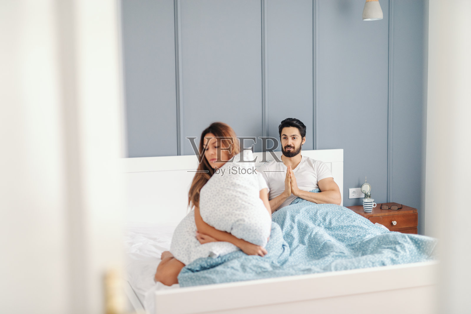 愤怒的女人坐在床上，抱着枕头背对着她的丈夫，丈夫在乞求她的原谅。卧室内。照片摄影图片