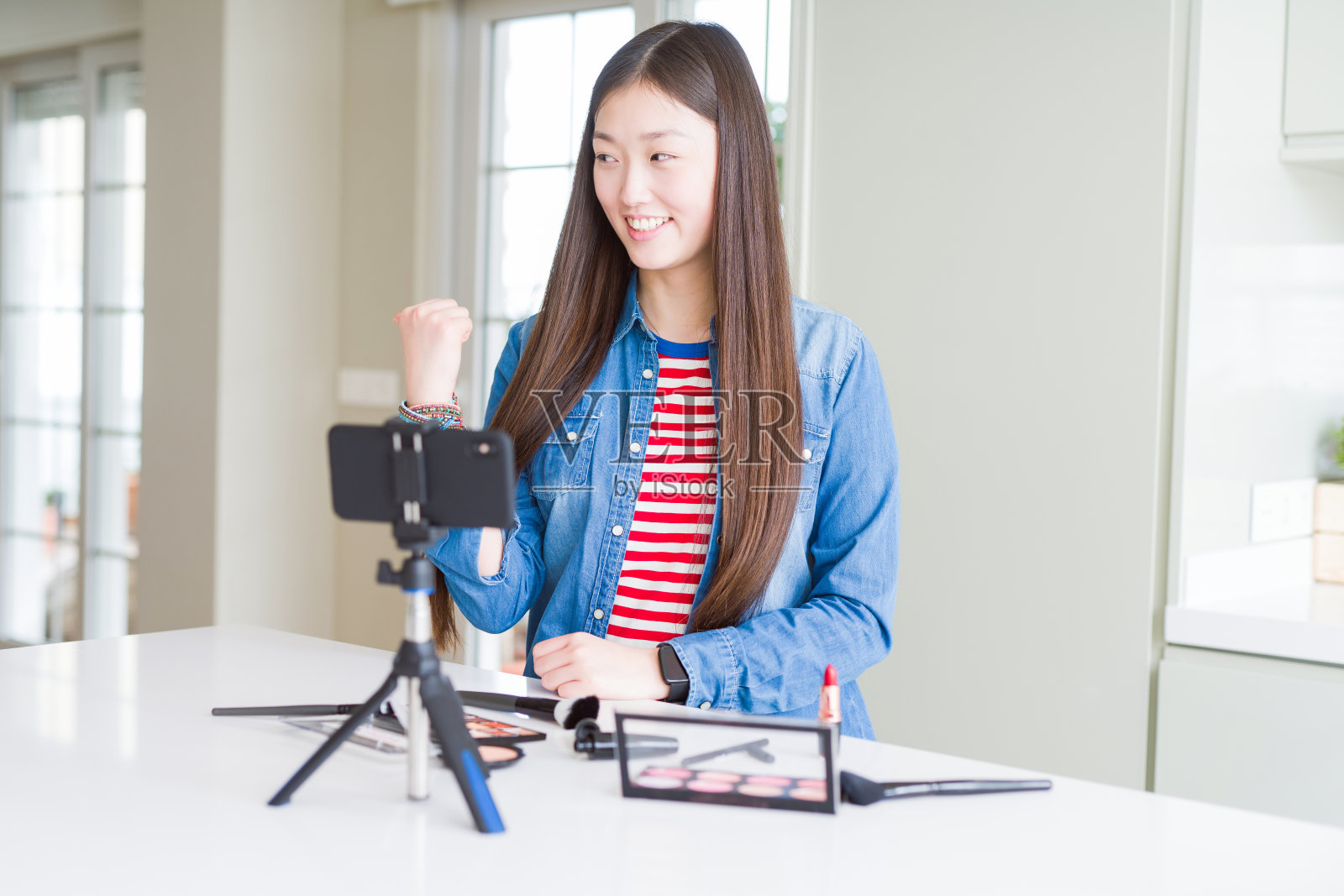 美丽的亚洲影响女人记录化妆教程使用智能手机摄像头微笑，快乐的脸看和指向一边的拇指向上。照片摄影图片