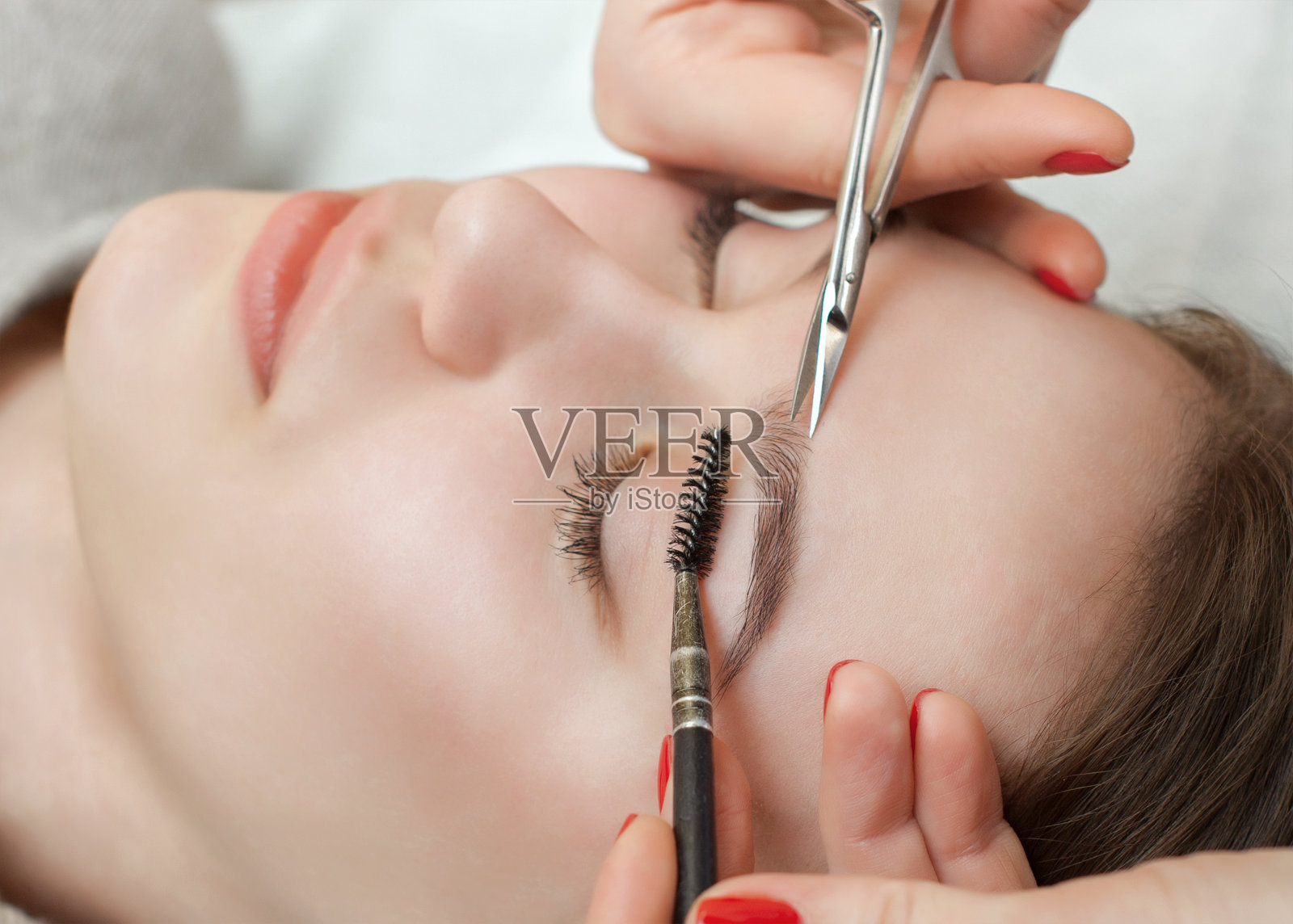 化妆师用剪刀剪她的眉毛。长着漂亮的长睫毛和修剪整齐的眉毛的女孩。照片摄影图片