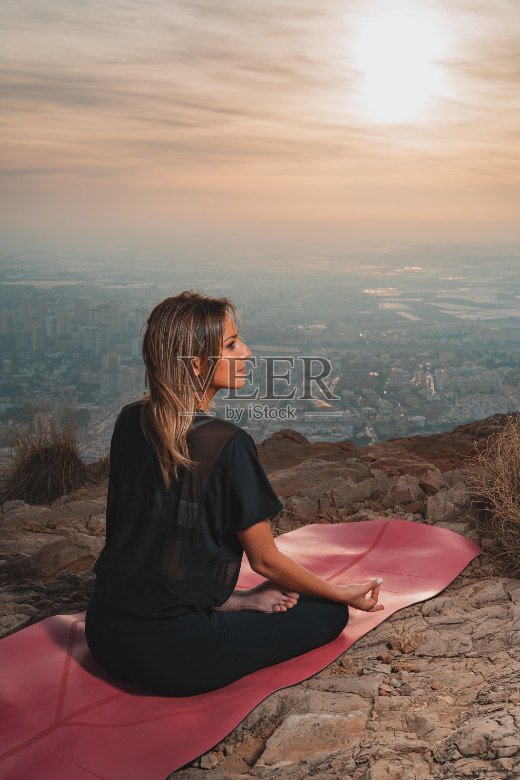 美丽的年轻女子在山顶做瑜伽，在美丽的夕阳下从城市看风景。自由和瑜伽概念。照片摄影图片