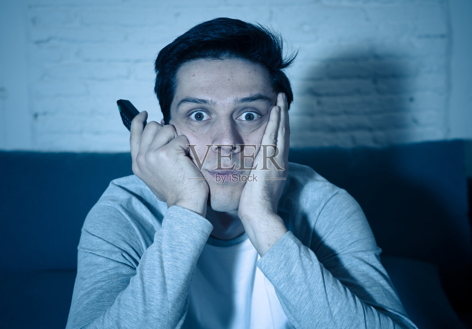 生活肖像的年轻人感到害怕和震惊，做出恐惧，焦虑的手势，同时看电视和拿着遥控器。暴力恐怖电影和系列网络流媒体。照片摄影图片