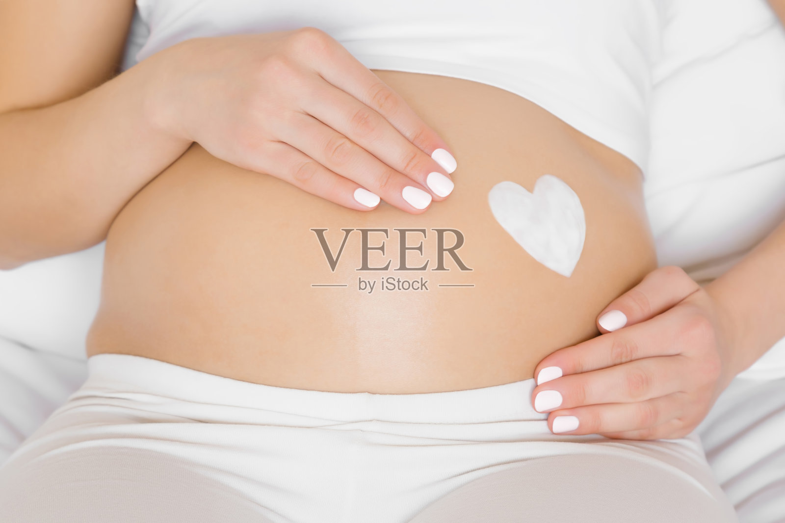 女孩坐在床上。年轻女子的手抚摸着她赤裸的大肚子。奶油做成的心形。呵护怀孕期间完美、柔软、光滑的肌肤。关闭了。前视图。照片摄影图片