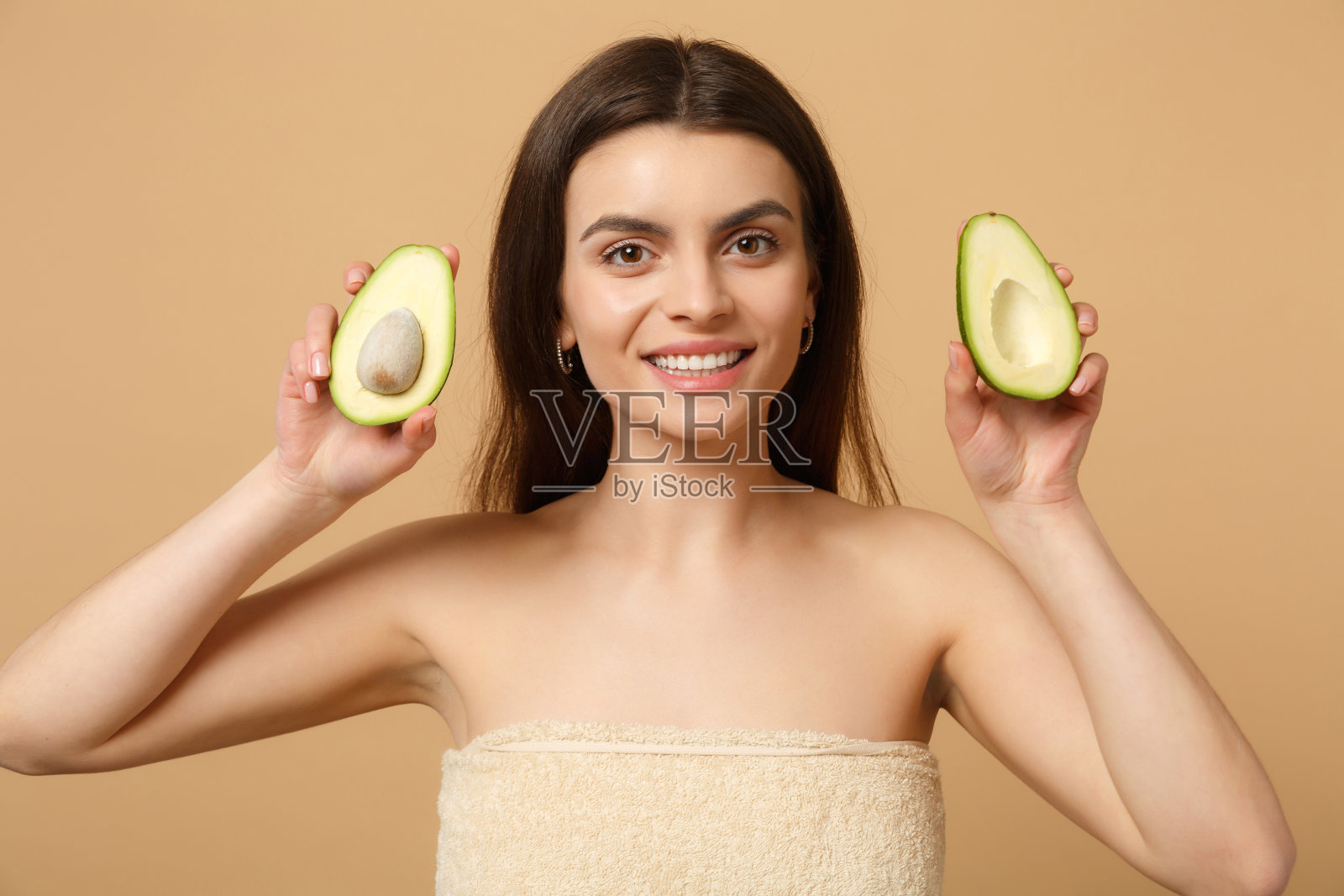 近距离黑发半裸女人20多岁与完美的皮肤化妆持有鳄梨孤立的米色粉彩墙背景工作室肖像。保健美容程序概念。模拟拷贝空间。照片摄影图片