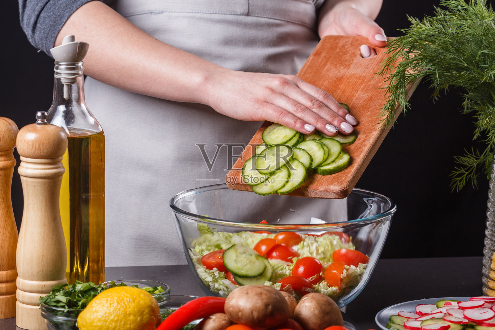 一位身穿灰色围裙的年轻女子在沙拉中加入了黄瓜照片摄影图片